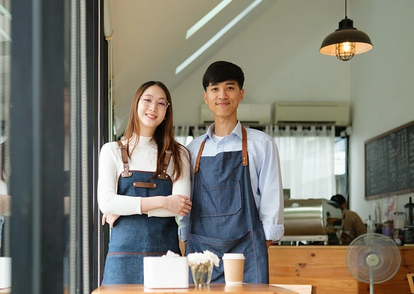 A smiling man and woman in aprons stand in a cozy café with wood tables. The atmosphere is warm and inviting, with natural light and a modern design.