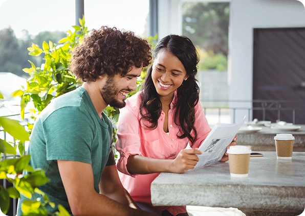 A smiling woman and man sit at a cafe table, with the woman holding and showing a paper to the man. Two coffee cups are on the table. Bright and cheerful.