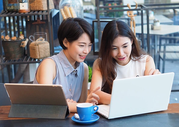 Two women are smiling and looking at a laptop in a cozy cafe. A tablet, blue coffee cup, and rustic shelves are visible, conveying a collaborative and relaxed atmosphere.